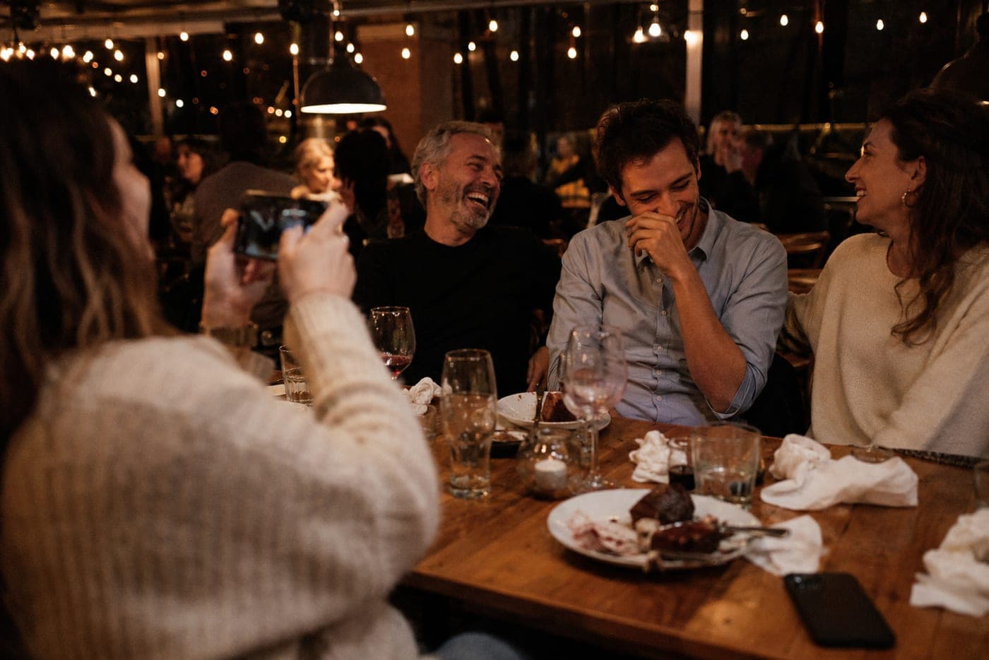 Colleagues laughing together at a company dinner while one of them photographs the moment on her phone.